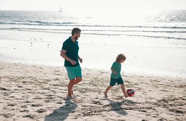 Father and son play soccer or football on the beach on summer family holidays. Dad and child playing outdoor