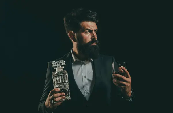 Barman or Bartender serves cognac. Bearded handsome man holding glass of whiskey. Confident bearded man in black suit with glass of whisky in loft