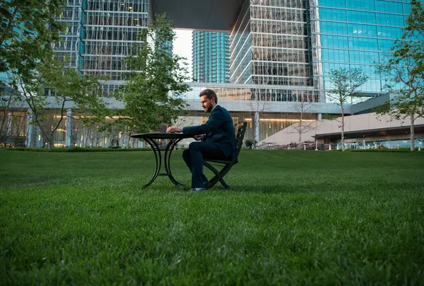 Businessman on large grass field near modern office building. Businessman with laptop in park of megapolice