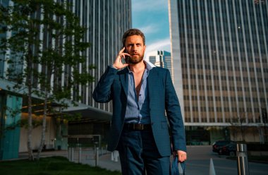 Şehirde yürüyen bir iş adamı. Şehir merkezindeki ofisin dışında cep telefonu kullanan bir iş adamı. Arkasında gökdelenler olan binalar. Beyaz adam iş için akıllı telefon tutuyor.