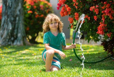 Çocuk bahçede bahçe hortumuyla oynuyor. Komik heyecanlı çocuk arka planda su püskürterek eğleniyor. Yaz çocukları açık hava aktivitesi