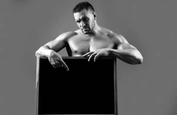 Handsome bodybuilder man with sign advertising board. Strong muscular athlete holding blank black poster ad. Copyspace