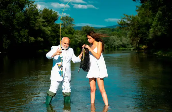Old senior and girl fishing. Young sexy woman and old fisherman standing in river with fishing rod. Father and daughter fishing. Mature rich man fisher celebrate retirement