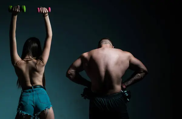 Muscular man with naked body, fitness woman with dumbbells on a dark background, back view. Sexy sport couple exercising with dumbbell. Slim and healthy sexy girl with strong muscular man workout