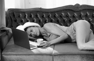 Dizüstü bilgisayarı ve pijamalı kahvesi olan güzel, şehvetli genç bir kadın. Yatak odasındaki boş vakitlerin tadını çıkarıyor.