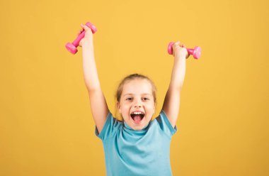 Child girl is doing exercises with dumbbells on yellow studio background. Sport portrait kids