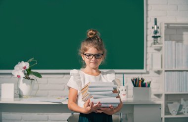 Cute child at school. Kid is learning in class on background of blackboard. Nerd school girl in glasses with books on blackboard