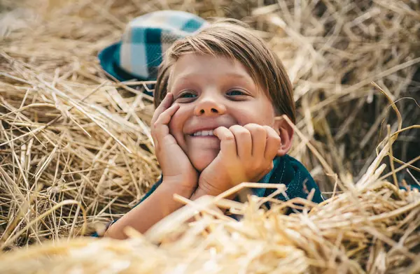 干し草の中で頭に帽子をかぶった喜びに満ちた男 その少年は秋に子供服を宣伝する 幸せな子供時代村の秋の休日にかわいい男の子 — ストック写真