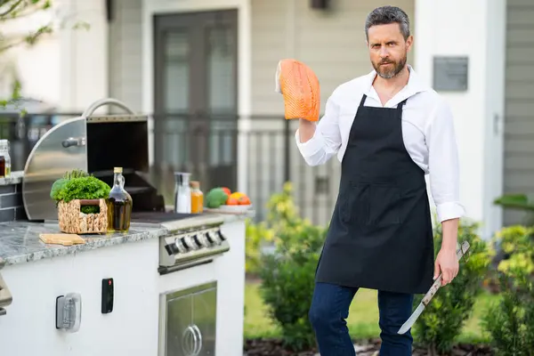 在房子的后院 穿着围裙做饭的男人在烤肉上烤鲑鱼鱼 一个英俊的男人在准备鲑鱼烧烤烤肉主厨烤肉和烧烤 — 图库照片