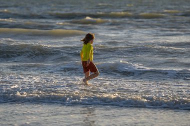 Heyecanlı çocuk yaz günü tropikal plajda koşup oynuyor. Çocuk dışarıda eğleniyor. Denizde yaz tatili. Gün batımında sahilde koşan küçük bir çocuk. Yaz tatilindeki çocuklar