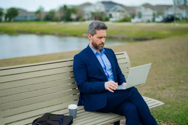 Business man sitting on a bench in park. Man is working on computer outside. Handsome business man making online work from laptop sitting on park bench. Business man in suit reading news in park