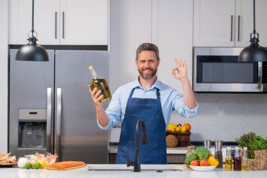 Mutfakta zeytin yağıyla yemek hazırlayan önlüklü bir adamın portresi. Şef önlüklü yakışıklı adam, zeytinyağıyla tutun. Aşçı mutfakta yemek yapıyor. Milenyum adamı zeytinyağı şişesi tutuyor.