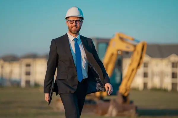 Successful construction business owner. Construction worker in suit and helmet near excavator. Confident construction owner in front of construction site. Civil engineer worker