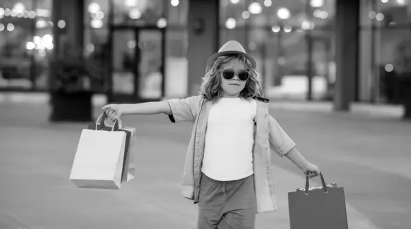 夏のファッションの服でかわいい男の子は買い物に行きます 手にショッピングパッケージと幸せな子供 買い物袋の屋外で買い物をする子供 買い物袋を持って通りを歩くかわいい子供 — ストック写真
