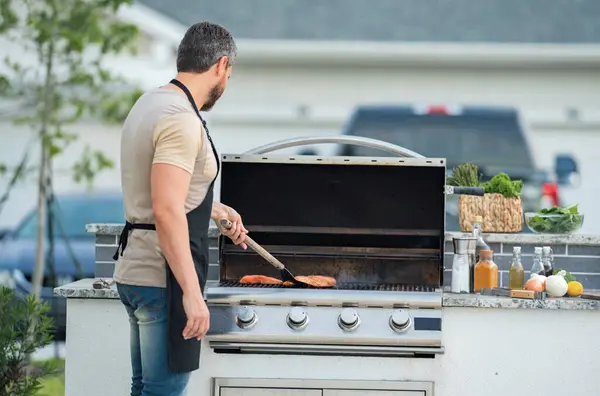 Man at barbecue grill. Male cook preparing barbecue outdoors. Bbq meat, grill for picnic. Roasted on barbecue. Man preparing barbeque in the house yard. Barbecue and grill. Cook using barbeque tongs