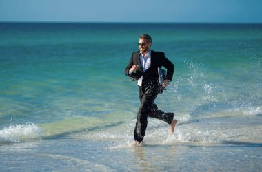 Leisure weekend and remote freelance work. Crazy comic business man in suit rest on sea bech. Funny businessman with laptop runnig on water. Remote summer business. Summer holiday for businessman