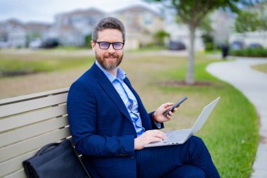 Takım elbiseli adam parkta dinleniyor. Bankta oturan bir iş adamı. CEO dışarıda dizüstü bilgisayarda çalışıyor. İnternette dizüstü bilgisayardan çalışan yakışıklı bir adam parkta bankta oturuyor. Takım elbiseli iş adamı dizüstü bilgisayar kullanmış.