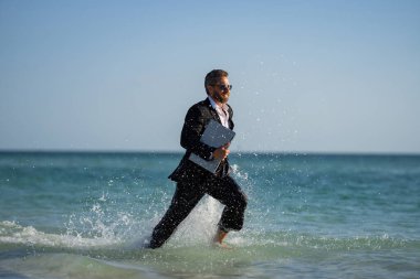 Yaz seyahati. İş başarısı. Serbest çalışma, uzaklık, online çalışma, e-çalışma. Takım elbiseli bir işadamı deniz suyunda dizüstü bilgisayarla çalışıyor. Komik bir yaz işi. Serbest seyahat.