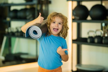 Heyecanlı çocuk jimnastik salonunda yoga minderi tutuyor. Yoga çocuk konsepti. Genç güçlü sportif çocuk kendini güçlü ve enerji dolu hissediyor. Spor sporu konsepti