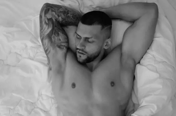 Seductive gay. Muscular body of sexy man in bed. Strong brutal guy. Sexy male naked torso. Nude muscular body man with tattoo lay in the bed. Muscular guy laying on bed