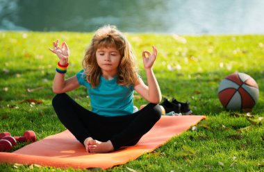 Çocuk yoga yapıyor, spor kıyafetleri giyiyor, dışarıda tişört giyiyor. Yoga sırasında meditasyon yapan çocuklar. Küçük çocuk nilüfer çiçeği pozunda meditasyon yapıyor. Jimnastik, çocuklar için meditasyon