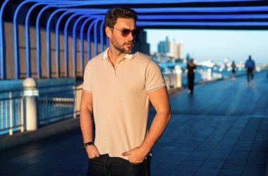 Sokakta polo tişörtlü yakışıklı bir genç. Dışarıda poz veren yakışıklı, kendine güvenen bir hippi modeli. Amerikan şehrinin arka planında seksi moda erkeği