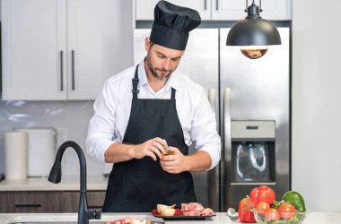 Mutfakta yakışıklı bir adam sağlıklı meyve salatası ve et hazırlıyor. Önlüklü bir adam ve aşçı şapkası mutfakta yemek hazırlıyor. Aşçı şapkası aşçısı mutfakta lezzetli yemekler pişiriyor. Modern mutfak iç mimarisi