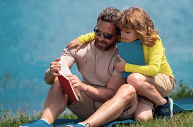Baba oğluyla parkta kitap okuyor, okul ödevlerini hazırlıyor, ebeveynlik yapıyor. Yaz yaşam tarzı. Ebeveynlik ve çocukluk kavramı. Küçük çocuk babasıyla açık bahçede öğreniyor.