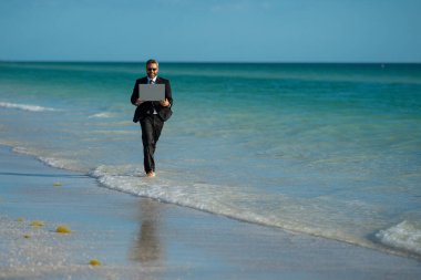 Serbest iş, online iş. Yaz işi. Takım elbiseli iş adamı dizüstü bilgisayarı tutuyor ve denizde koşuyor. Suda dizüstü bilgisayarı olan takım elbiseli komik bir iş adamı. Uzaktan kumanda. Çılgın serbest çalışan.