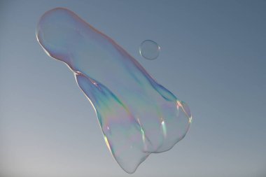 Mavi bulutlu gökyüzüne karşı havada büyük sabun baloncukları patlatıyorlar. Gökyüzünde yüzen büyük baloncuk, gökyüzü parlak mavi arkasında, kabarcık şeffaf ama spektrumun renklerini yansıtıyor