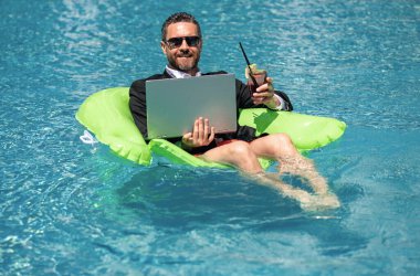 Serbest çalışma, uzaklık, online çalışma, e-çalışma. Yaz işi. Takım elbiseli bir iş adamı yaz kokteyli içer ve bilardoda dizüstü bilgisayar kullanır. İş adamı yazın havuz suyunda iş yapmayı hayal eder.