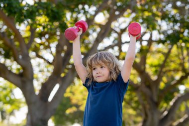 Yaz parkında çocuklar spor eğitimi alıyor. Çocuk spor egzersizleri. Sağlıklı çocuk hayatı ve spor anlayışı. Dambıllarla çalışan çocuk portresi. Çocuklar için motivasyon ve spor kavramı