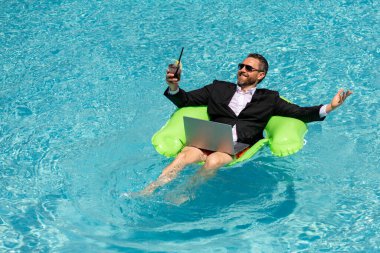 Takım elbiseli komik iş adamı yaz kokteyli içer ve deniz sahilinde dizüstü bilgisayar kullanır. Yaz tatili, açık hava ve yaz seyahati konsepti. Çılgın ofis çalışanı yazın havuz başında hayal kurar.