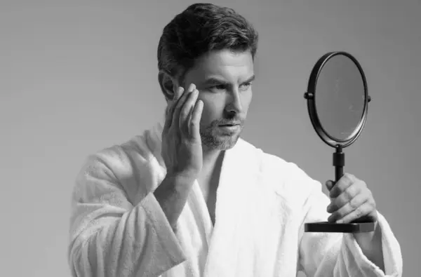 Man applying cosmetic face cream on mirror. Facial treatment. Portrait of man with bare naked shoulders touching skin. Sexy man with skin care product. Male face creme. Perfect skin, morning routine