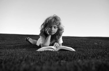 Yeşil çim tarlasında kitap okuyan sevimli çocuk. Akıllı çocuk kitap okuyor, arka planda çim parkında yatıyor. Zeki çocukların portresi.
