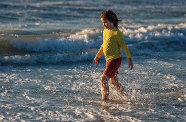 Güneşli bir yaz gününde ıslak sahilde koşan kaygısız küçük çocuk çıktı. Çocuk yaz plajında koşuyor. Tasasız çocuk. Heyecanlı çocuk yaz denizinde koşuyor. Tatlı çocuk yaz denizinde koşuyor.