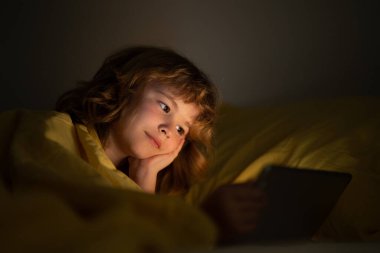 Çocuk tablet bağımlısı, çocuk tablet kullanıyor, çizgi film izliyor. Çocuk yatakta tabletini izliyor. Aygıt ekranından aydınlatılmış çocuk yüzü. Yorganın altındaki çocuk bir tablet tutuyor. Gece vakti.