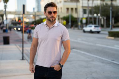 Sokakta polo tişörtlü yakışıklı bir genç. Dışarıda poz veren yakışıklı, kendine güvenen bir hippi modeli. Amerikan şehrinin arka planında seksi moda erkeği