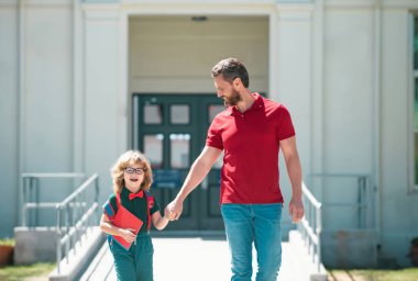 Parent and pupil of primary school go hand in hand. Teacher in t-shirt and cute schoolboy with backpack near school park clipart