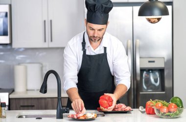 Önlüklü adam ve aşçı şapkalı adam mutfakta yemek pişiriyor. Mutfakta sağlıklı yemek pişiren yakışıklı adam. Adam mutfakta yemek pişiriyor. Taze yemek malzemeleriyle ev menüsü. Modern mutfak iç mimarisi