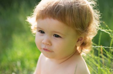 Doğada yeşil çimlerin üzerinde oynayan küçük bir bebek. Çocuklar oynuyor. Bebek yüzlü, yakın çekim. Küçük komik çocuk portresi. Sarışın çocuk, duygusal yüz. Bebek ve yaz güneşli hava
