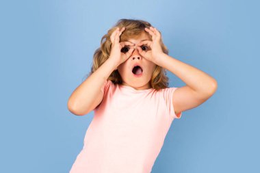 Heyecanlı çocuk parmaklarını gözlük gibi gözünün yanında tutuyor. Stüdyonun izole edilmiş arka planında dürbün gibi hareket eden çocuk.