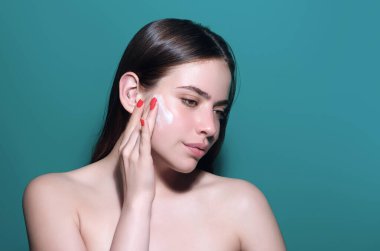 Beautiful model applying cosmetic cream treatment on face. Facial treatment. Beauty portrait of young topless woman with bare shoulders applying face cream