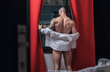 Handsome nude man in a bedroom. Young sexy man at night. Muscular handsome man in hotel room on window curtains. Shirtless topless sexy male model posing indoor. Sexy naked body