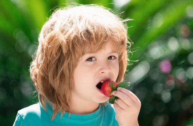 Sağlıklı çocuk yemekleri. Yeşil bahar arka planında çilek yiyen sevimli bir çocuk. Mutlu yüzlü çocukları kapatın.