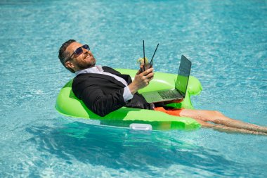 Yaz tatilleri ve özgürlük seyahati konsepti. Sahildeki komik iş adamının portresi. Takım elbiseli bir iş adamı dizüstü bilgisayar ve bilardoda kokteyl ile yüzüyor. Çılgın iş adamı yaz tatilinde rüya görür