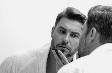 Adam aynaya kozmetik yüz kremi sürüyor. Yüz bakımı. Çıplak omuzlarıyla tenine dokunan bir adamın portresi. Cilt bakım ürünü olan seksi bir adam. Erkek yüz kremi. Mükemmel cilt, sabah rutini