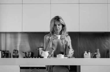 Güzel bir kadın mutfaktaki masanın yanında kahve içer. Büyük göğüsleri pijamayla kaplayan seksi manken. Ev hanımı sabah mutfakta.