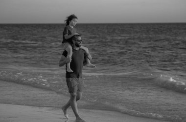 Baba ve oğul sahilde yürüyor. Yaz tatili. Baba ve oğul birlikte yaz tatilinin tadını çıkarıyorlar. Baba oğul sahilde oynuyorlar. Arkadaş canlısı bir aile. Oğullar babalarının omuzlarına biner.