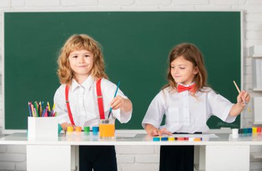 Sınıfta karatahta arka planda komik öğrenciler çiziyor. Okul arkadaşları çocuklar, kız ve erkek birlikte resim yapıyorlar. Sevimli çocuklar renkli pastel boyalarla resim çizerler.